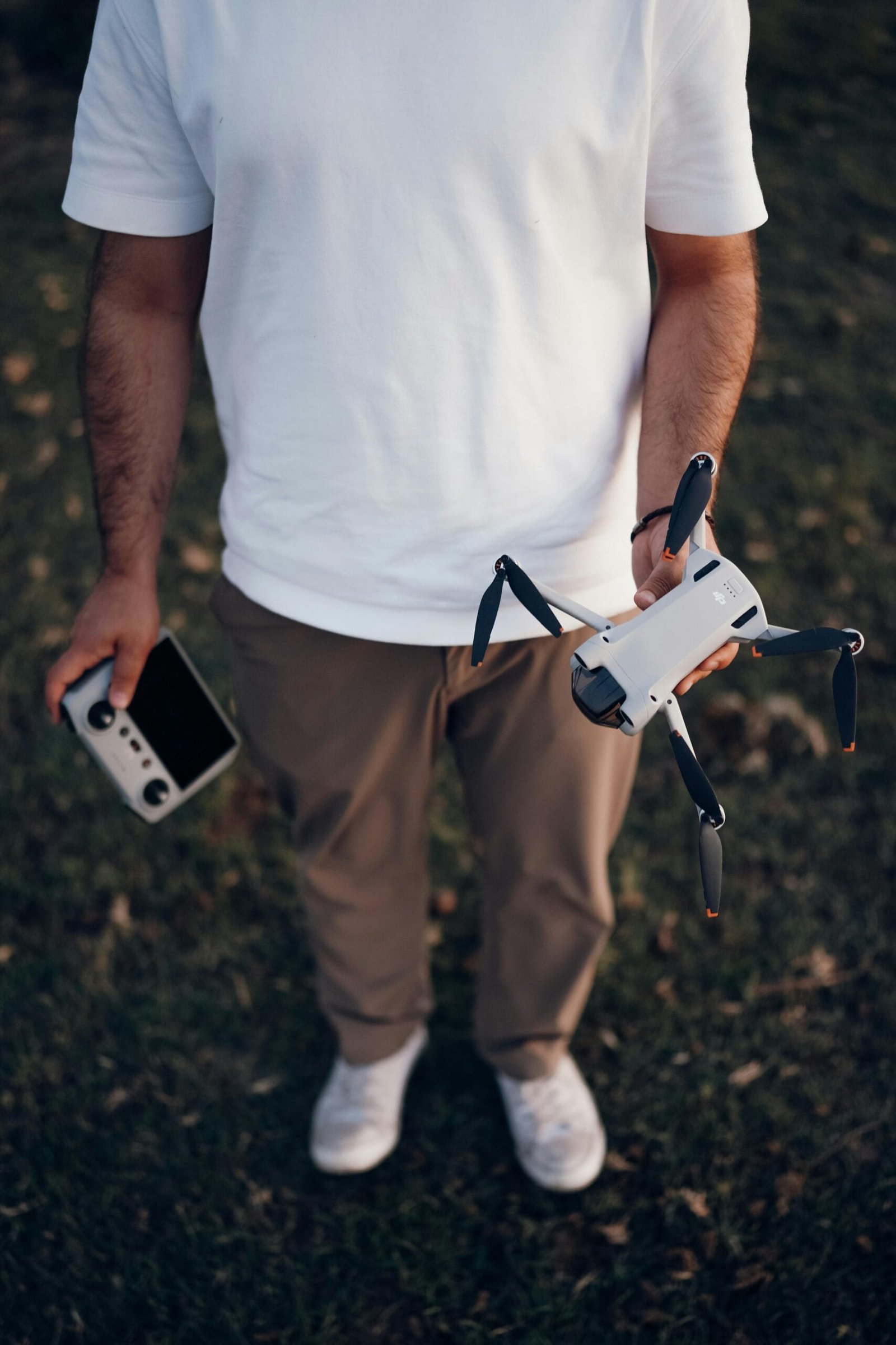 Man in white shirt holding a drone and remote controller on grass.